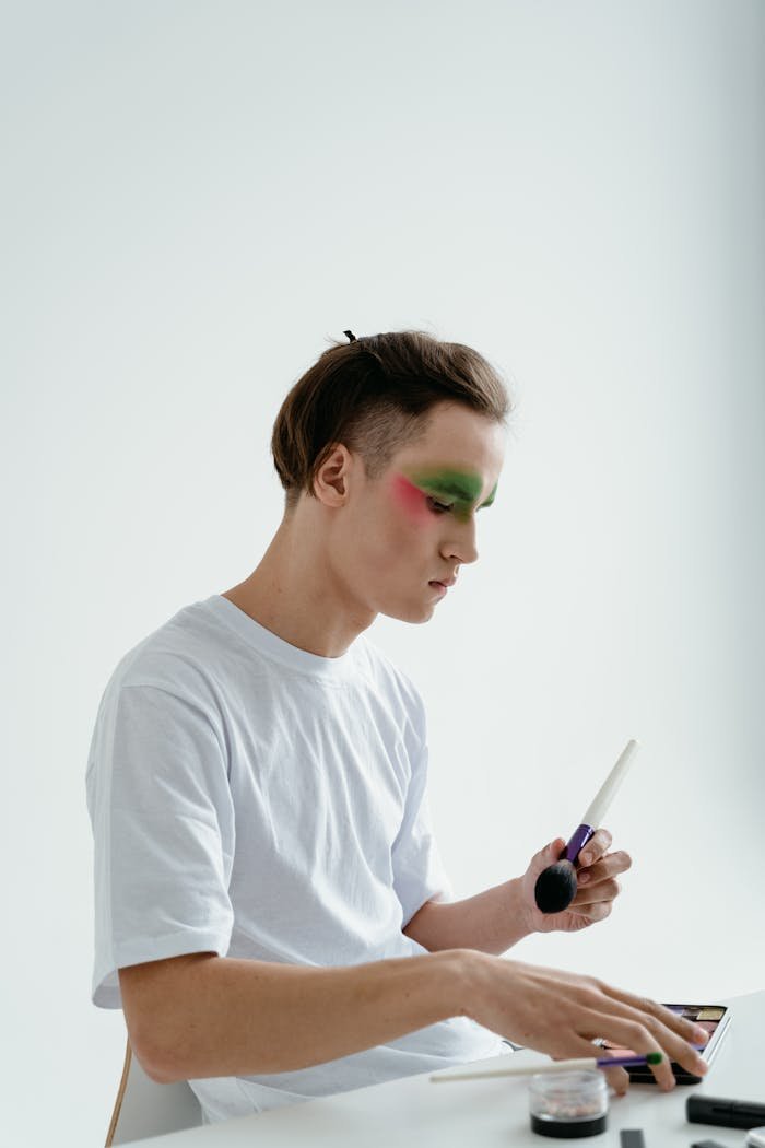 Young man using makeup brush to apply colorful eyeshadow in an indoor setting.