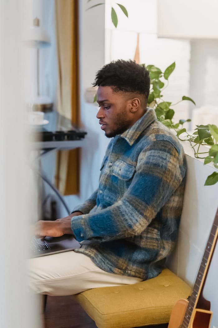 Side view of focused bearded African American guy in checkered shirt sitting near white partition decorated with green plant and working remotely on portable computer